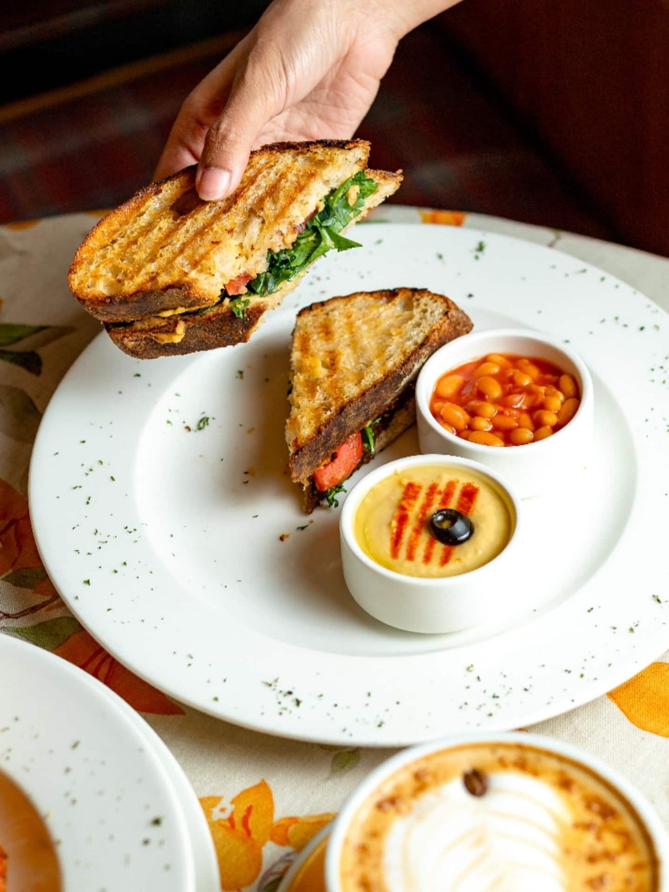 A grilled sandwich lifted from a plate with baked beans and a dip, a cappuccino beside it.