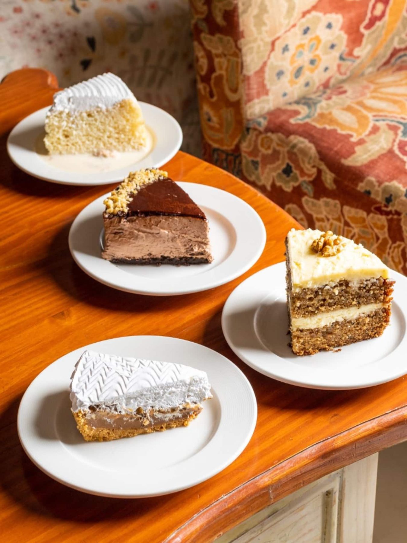 Four plated cakes on a wooden table at Café De Piccolo, a floral armchair behind.
