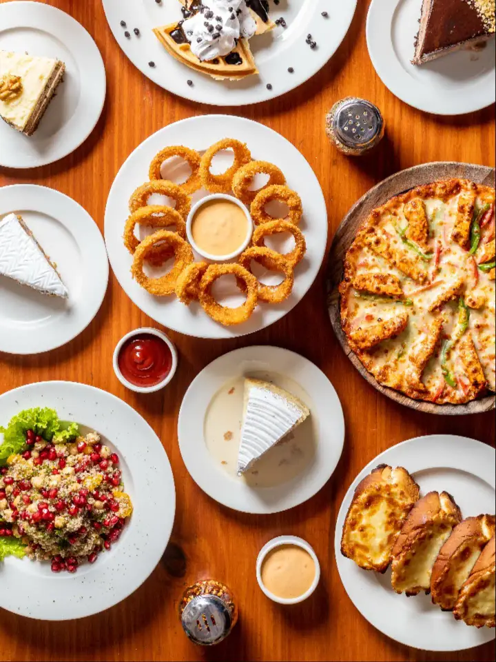 A table seen from above: a pizza, onion rings, a waffle, cakes, a salad and bread on white plates on an orange table.