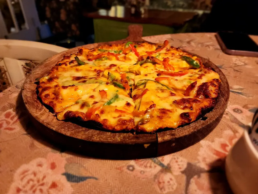 A pizza on a round wooden board, on a floral tablecloth, as a guest photographed it.