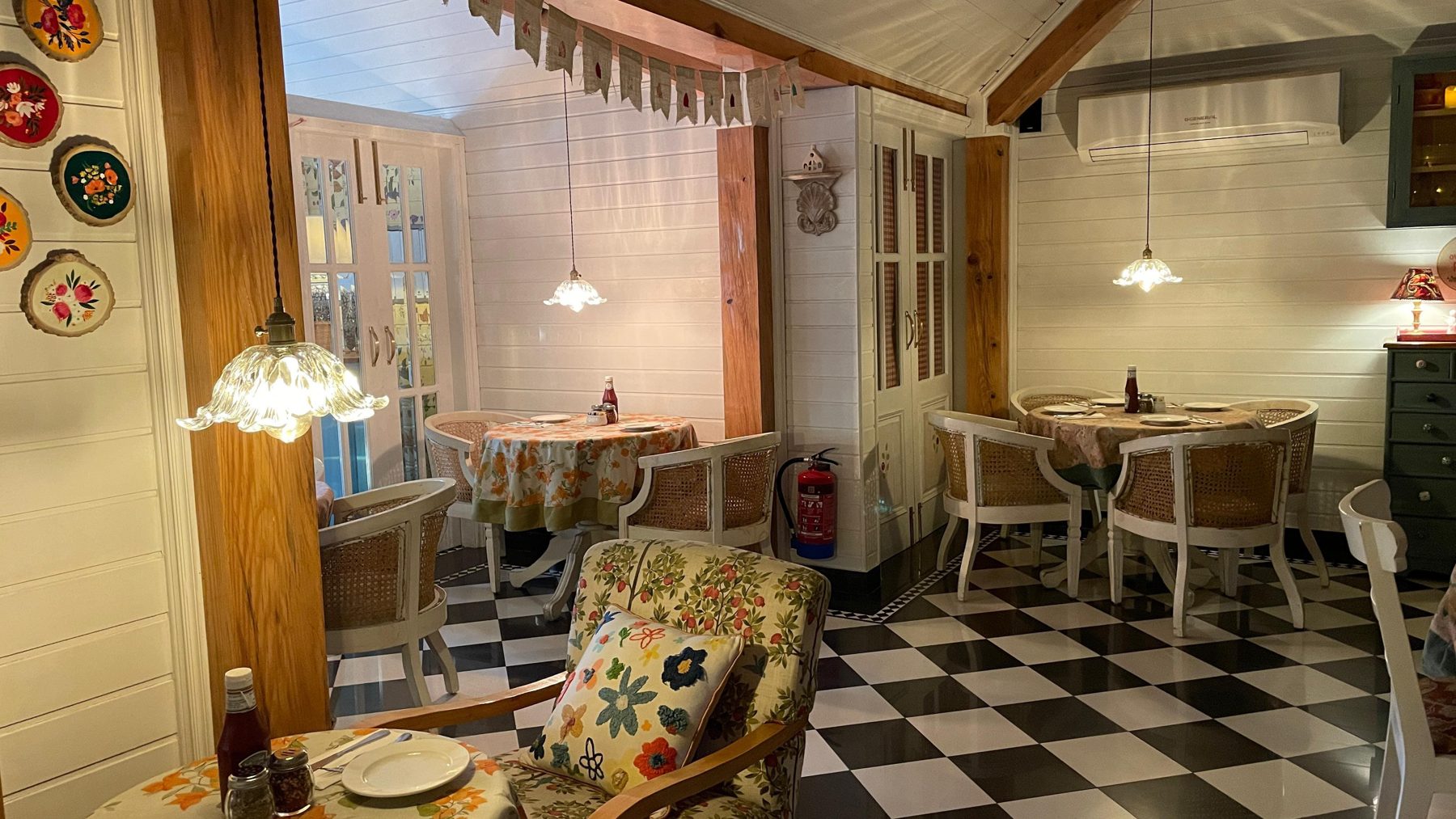 The room at Café De Piccolo: white plank walls, a checkerboard floor, cane chairs at set tables, a lamp on each.